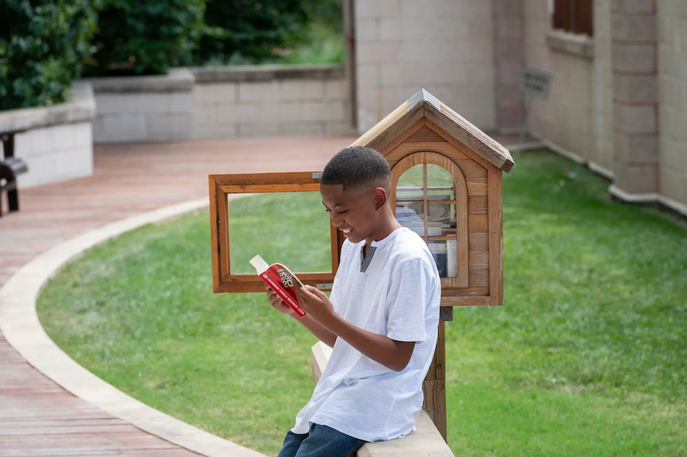 a boy leaning on a ledge, reading a book next to a little free library